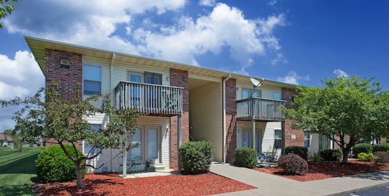 Two-story apartment building with brick and siding exterior, private balconies and patios, and landscaped grounds with trimmed shrubs, trees, and mulch beds