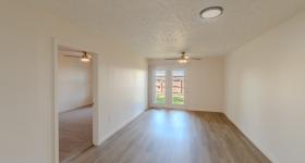 Spacious living room with wood-style flooring, neutral walls, and modern light fixtures. French doors open to a fenced outdoor area, and an adjoining doorway leads into the bedroom.