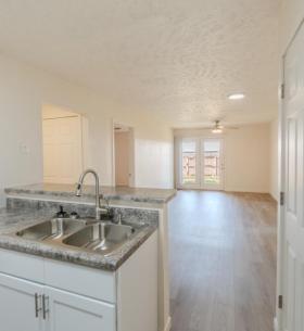 Spacious living room with wood-style flooring, neutral walls, and modern light fixtures. French doors open to a fenced outdoor area, and an adjoining doorway leads into the bedroom.