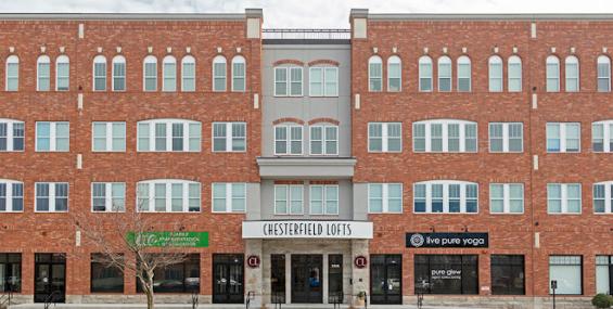 A wide shot of Chesterfield Lofts, highlighting its symmetrical brick architecture with large windows and decorative detailing. The ground floor features multiple businesses, including a yoga studio and wellness center, with additional commercial spaces available for lease. The entrance is centrally located, flanked by retail spaces designed for high foot traffic.
