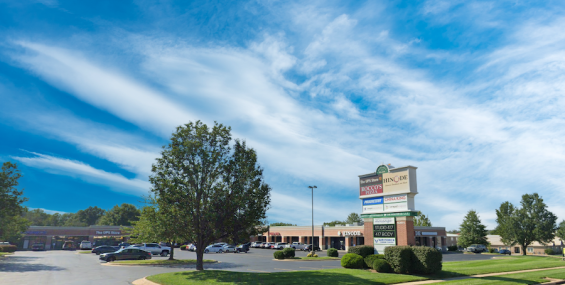 A wide-angle view of Aundria Plaza, a shopping center with a large parking lot in the foreground and a tall business directory sign. The sign displays several business names, including The UPS Store, Rocco’s Pizza, Hinode Japanese Steakhouse, China King, and others. The plaza is surrounded by green landscaping, and the sky above is bright blue with wispy white clouds.