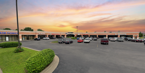 A closer view of Aundria Plaza at sunset, showing a row of brick storefronts with large glass windows. Businesses such as The UPS Store, Rocco’s Pizza, and Hinode are visible. The parking lot has several cars parked, and a few are driving through. The sky behind the plaza is filled with warm orange and pink hues.