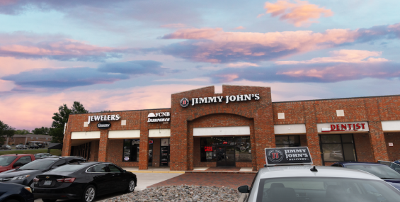 A brick strip mall features several businesses, including &quot;Jimmy John’s,&quot; a jewelry store, an insurance office, and a dentist. A white Jimmy John’s delivery car is parked in front, along with several other vehicles. The parking lot has a decorative rock bed. The sky in the background is a blend of warm pink and blue colors at dusk.