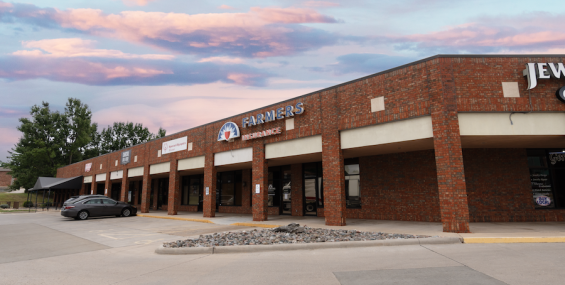 A brick shopping center with covered walkways and multiple storefronts, including &quot;Farmers Insurance,&quot; is shown. A silver sedan is parked near the entrance, and additional businesses are visible in the distance. The sky is filled with pink and blue tones at sunset, casting a warm glow over the scene.