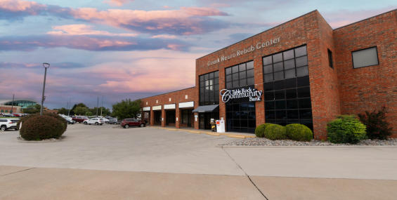 A brick commercial building with large glass windows houses &quot;Table Rock Community Bank&quot; and &quot;Ozark Neuro Rehab Center.&quot; The parking lot is mostly empty, with a few cars visible in the distance. Landscaping includes trimmed bushes and rock beds. The sky is a mix of pink and blue hues, indicating sunset.
