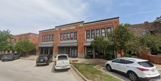 A two-story red brick office building with large windows and a classic architectural design. A sign above the entrance reads &quot;Dearborn Office Building.&quot; The facade features metal awnings over the ground-floor entrances, and trees line the front. Several cars are parked along the curb.
