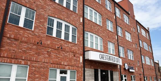 side-angle view of Chesterfield Lofts showcasing its curved brick facade and multiple floors of large, arched windows. The main entrance features a prominent &quot;Chesterfield Lofts&quot; sign above double glass doors. The building's ground-level commercial spaces offer a mix of storefront opportunities in a high-visibility area.
