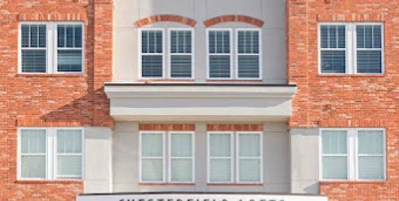 The front entrance of Chesterfield Lofts, a four-story brick building with large windows and a modern design. A sign above the entrance reads &quot;Chesterfield Lofts,&quot; and the ground floor features commercial spaces with glass storefronts. The exterior combines red brick with stone accents, creating a contemporary yet classic aesthetic.