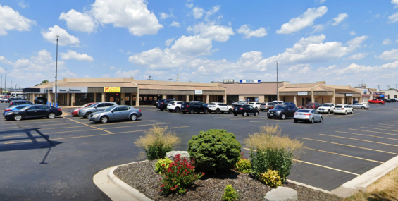 A shopping center with a row of tan-colored, single-story retail buildings under a bright blue sky with scattered white clouds. Visible businesses include a pharmacy, a chicken restaurant, and other retail stores. The large parking lot is partially filled with cars, and small, well-maintained shrubs and flowers line the edges of the lot. The shopping center appears well-maintained with clear signage.