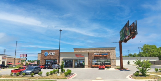 A modern shopping center with a stone and wood exterior, featuring AT&amp;T, Missouri Title Loans, and Mercy GoHealth Urgent Care. The storefronts have large glass windows, with visible signage for each business. A parking lot with cars and neatly trimmed landscaping is in front of the building. A tall billboard stands to the right, and a Planet Fitness sign is visible in the background. The sky is bright blue with a few scattered clouds, and the overall setting appears well-maintained and inviting.