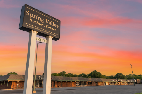  A tall, black and white sign reading "Spring Valley Business Center" stands against a colorful sunset sky. Below, the sign states that the center is professionally managed by The Wooten Co. LLC. The number "1675" is displayed on a separate panel beneath the main sign. In the background, a strip mall with brick storefronts and green trim lines an empty parking lot.